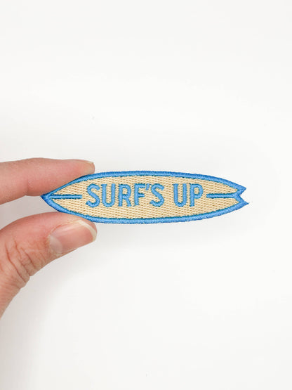 Hand holding a 'Surf's Up' patch against a white background