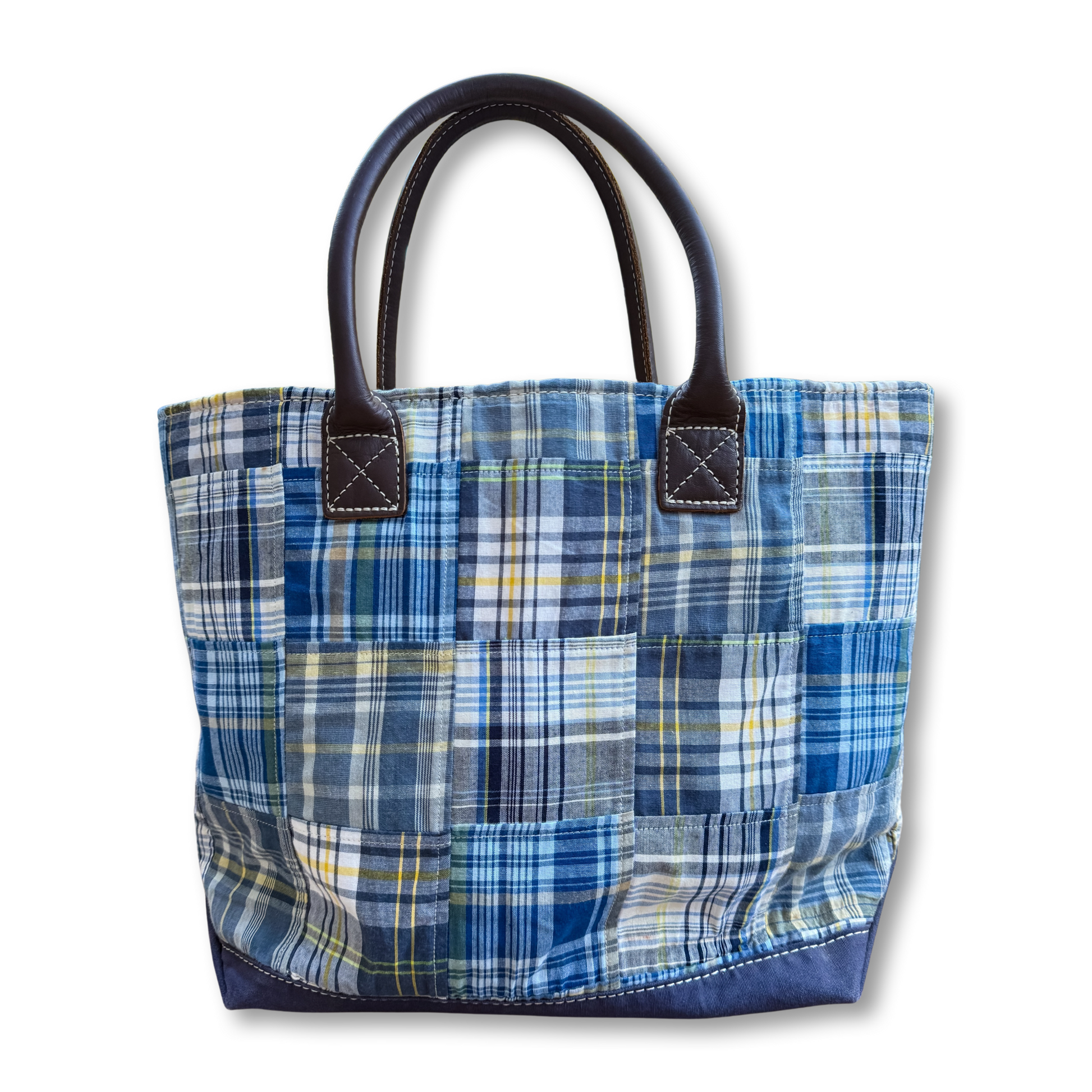 A handmade patchwork tote bag made from blue, navy, and white plaid fabrics, featuring dark brown leather handles and a solid navy blue base, shown against a plain white background.