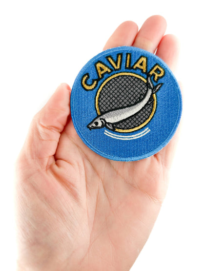 Circular patch with 'Caviar' text and fish illustration held in a hand against a white background