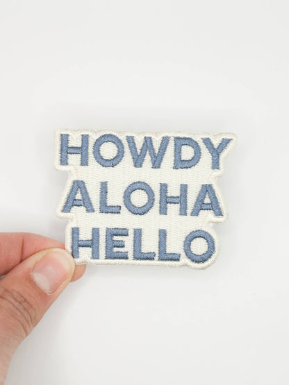 Embroidered patch with 'HOWDY ALOHA HELLO' text held by a hand on a white background