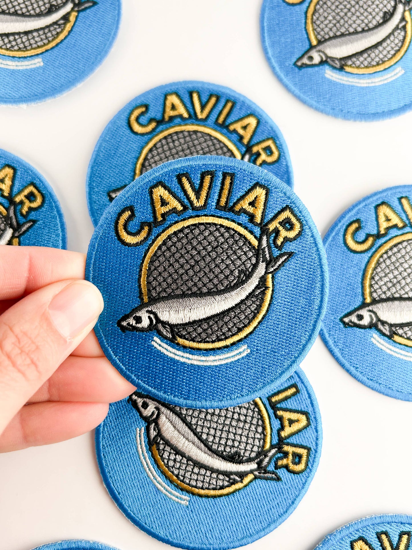 Blue caviar-themed patches with a hand holding one, on a white background.