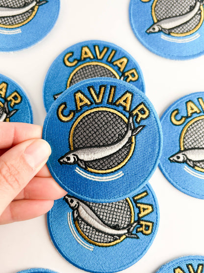 Blue caviar-themed patches with a hand holding one, on a white background.