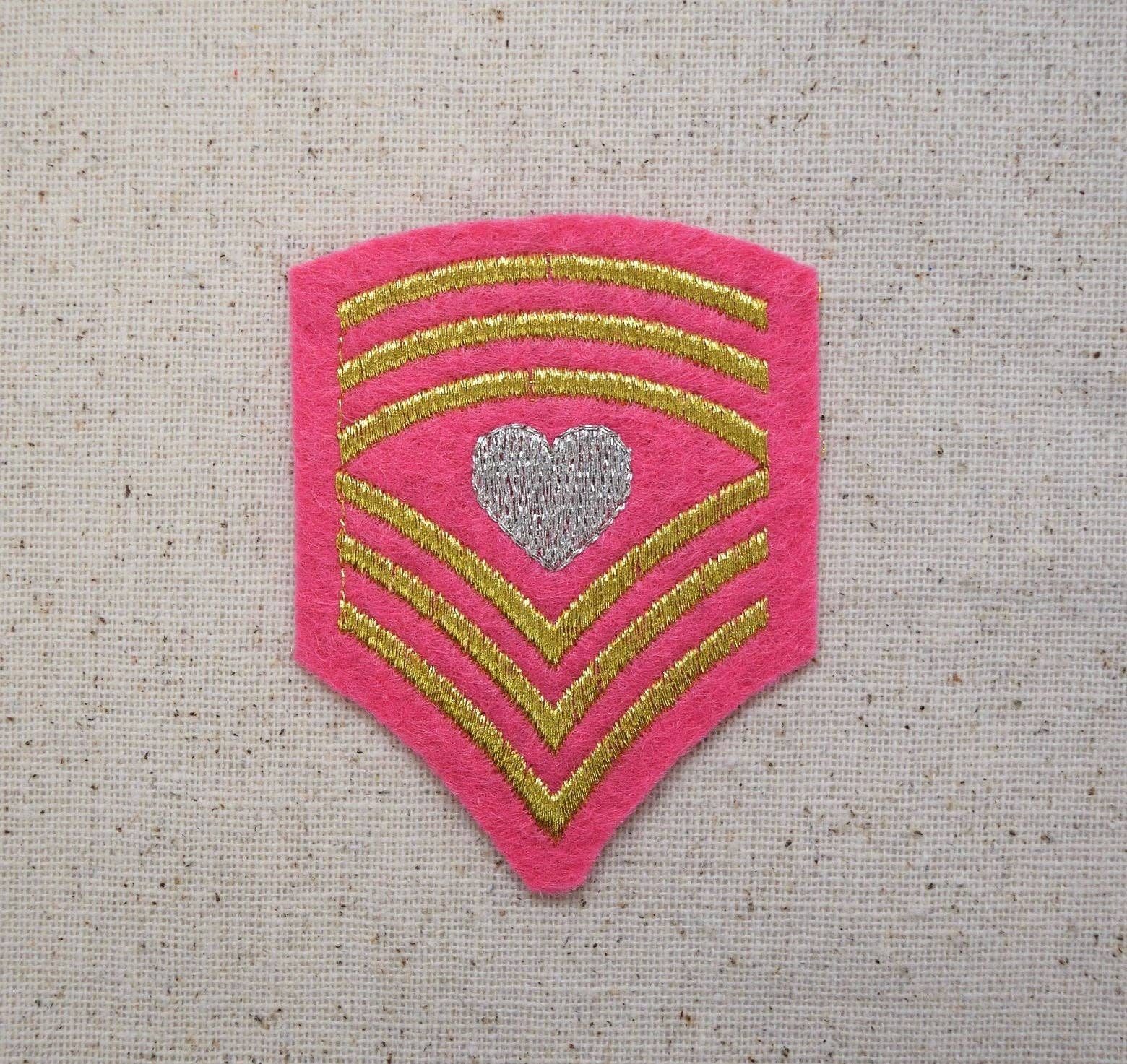 Pink military rank insignia shield patch with gold stripes and a heart emblem on a textured beige background