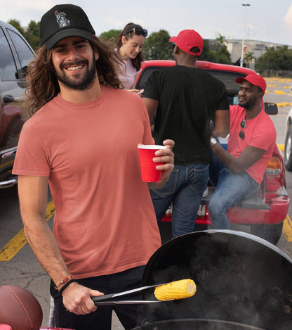 Man grilling corn on the cob in a parking lot with people and cars in the background.