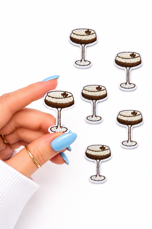 A hand with long light blue nails holds an embroidered patch of a champagne coupe glass, with five matching patches scattered on a white background.