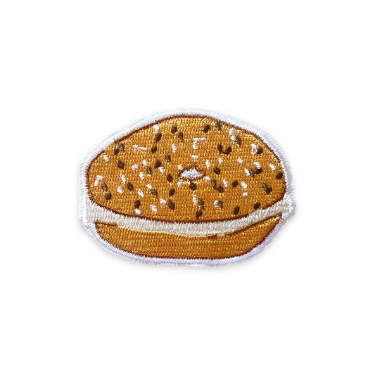 An embroidered patch shaped like a sesame seed bun, featuring a golden-brown top and bottom with white sesame seeds and a white border, shown on a plain white background.