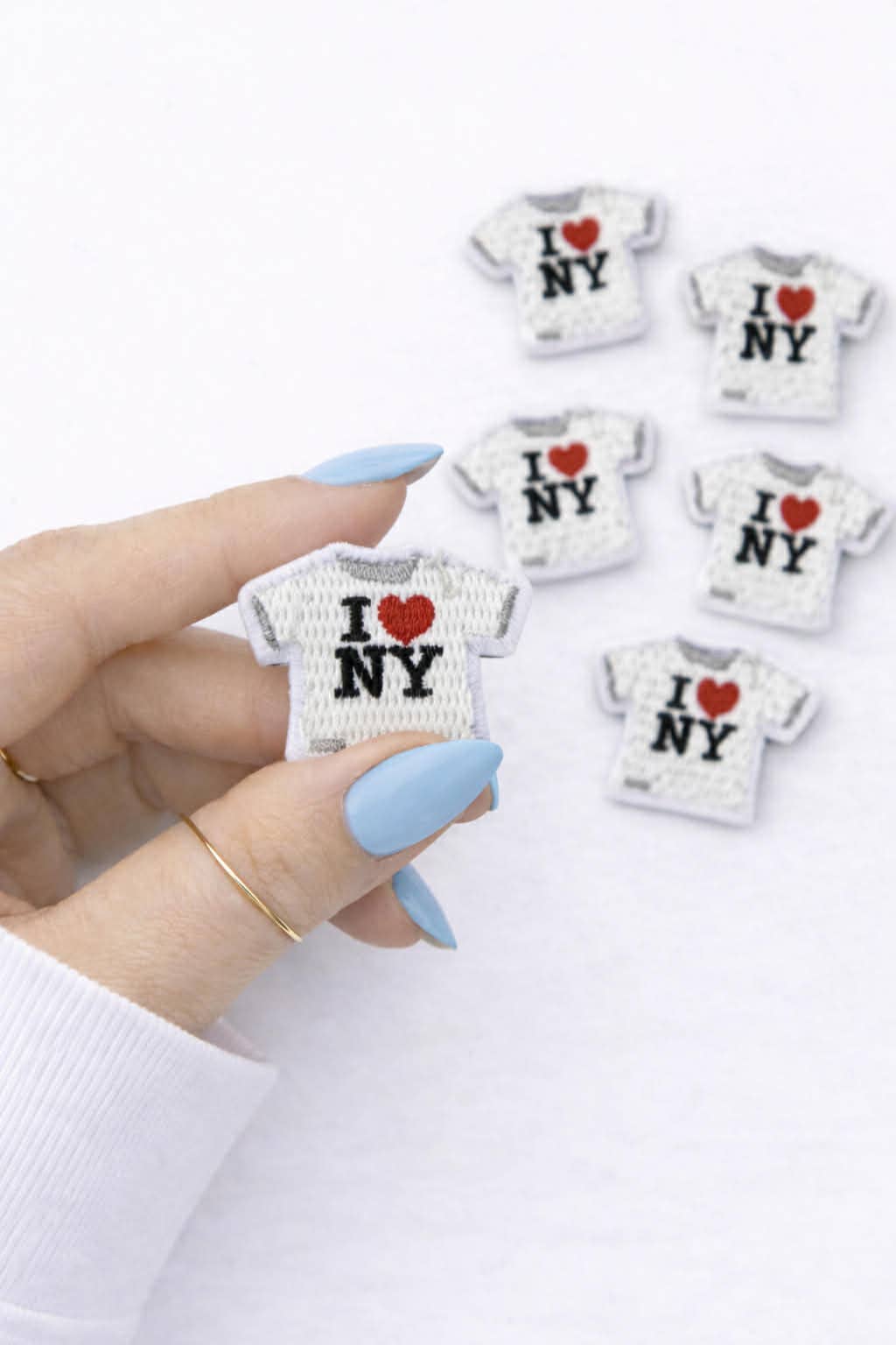Hand holding a miniature 'I Love NY' t-shirt with more t-shirts in the background on a white surface.
