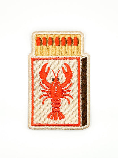 An embroidered patch shaped like a matchbox, featuring a red lobster design on the front, red matches at the top, and a dark brown side edge, shown on a plain white background.