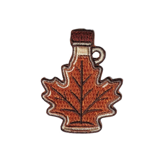 An embroidered iron-on patch shaped like a maple leaf, styled as a bottle with a brown cap and handle, featuring orange-brown, brown, and beige colors on a white background.