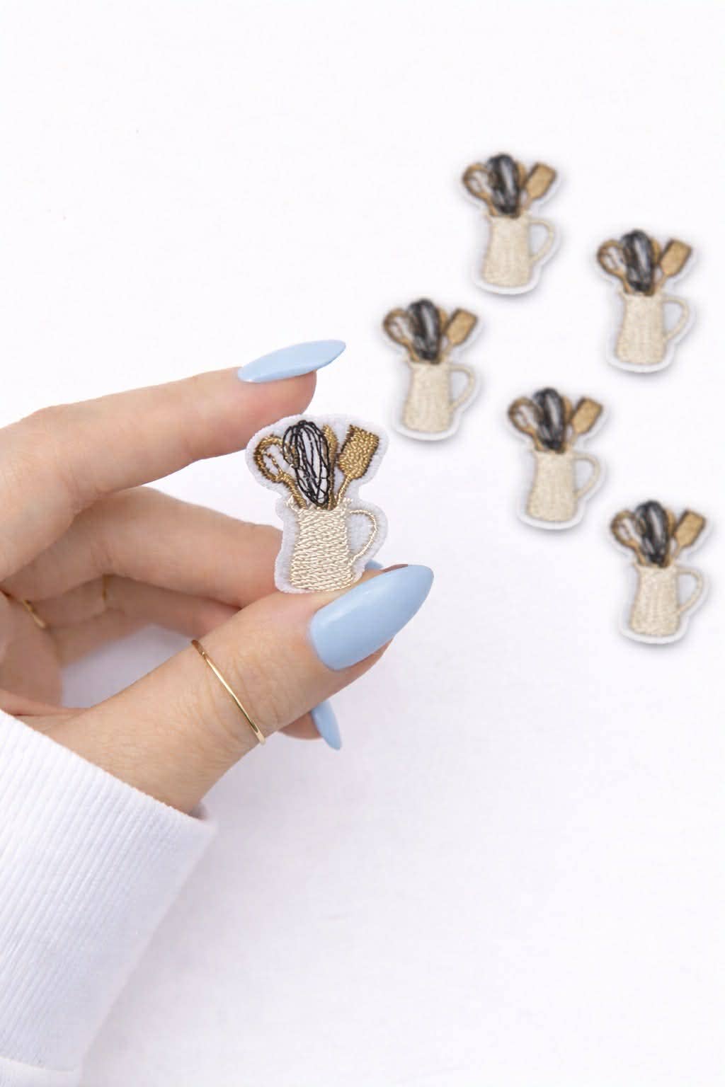 Hand with pale blue almond nails and thin gold rings holding a mini embroidered patch of a cream pitcher filled with a black whisk and gold wooden utensils, with five matching patches on a white background.