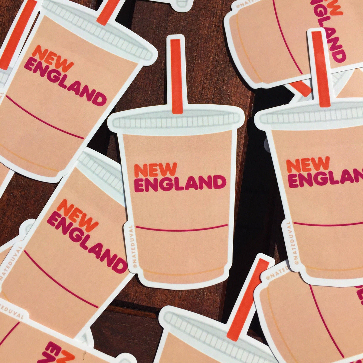 A collection of die-cut stickers shaped like takeaway coffee cups, scattered on a dark wooden surface. Each cup sticker is beige with the words 'NEW ENGLAND' in red and orange, with a red straw and white outline.