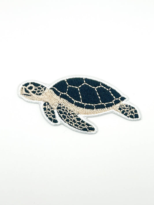 An embroidered sea turtle patch with a dark navy blue shell and head, cream-colored flippers and underside, and a white stitched border, shown on a plain white background.