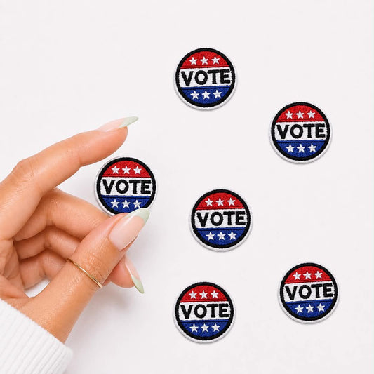 A hand with manicured nails holds one round embroidered patch while five identical patches are scattered on a plain white background. Each patch is red, white, and blue with white stars and the word VOTE in white text.